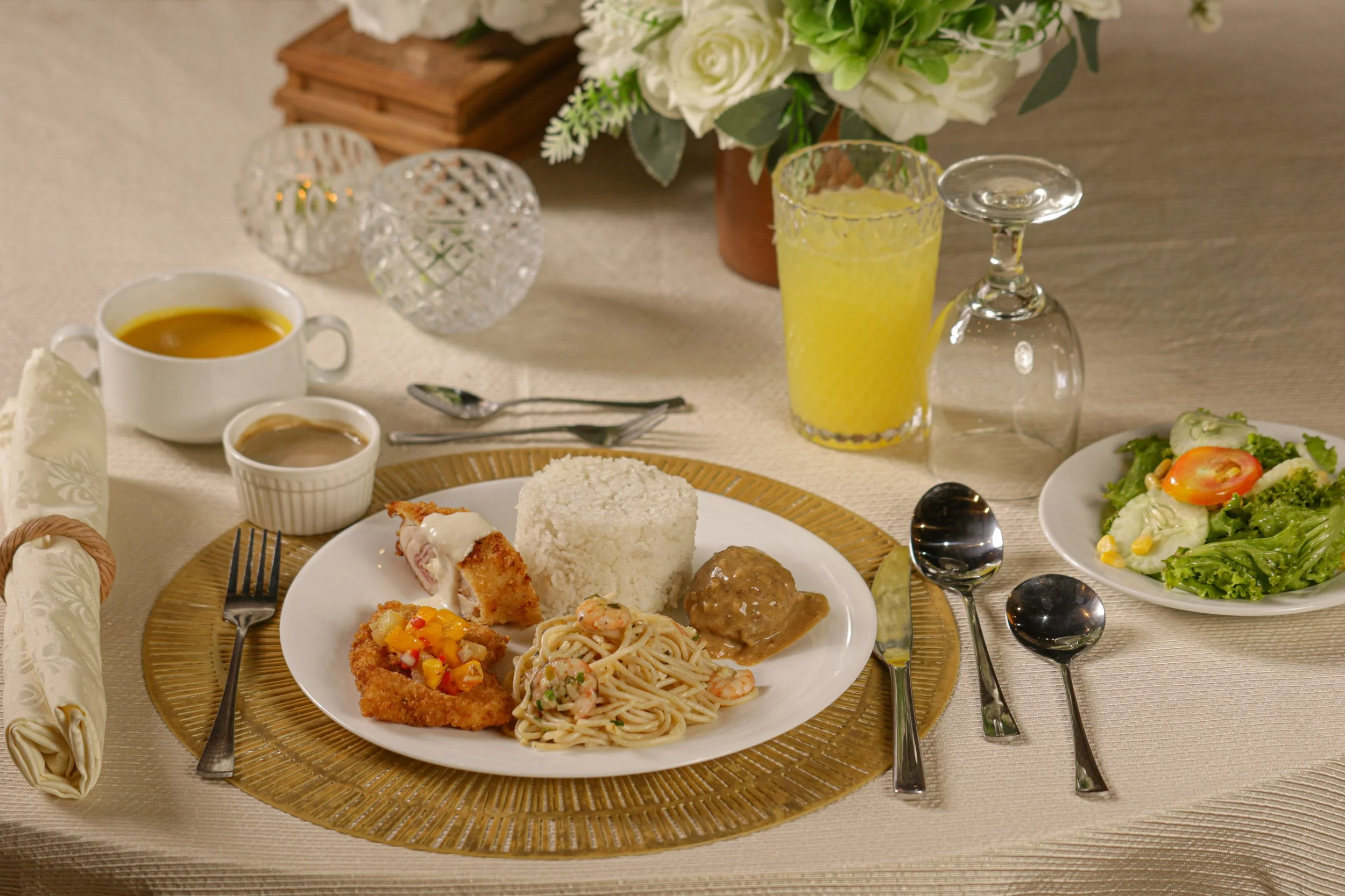 A dining table with a plate of rice, fried chicken with vegetables, and spaghetti with shrimp. There are two bowls of sauce, a glass of yellow drink, an upside-down wine glass, and a small side salad. The table is decorated with a floral centerpiece, crystal candle holders, and a rolled napkin with a napkin ring.