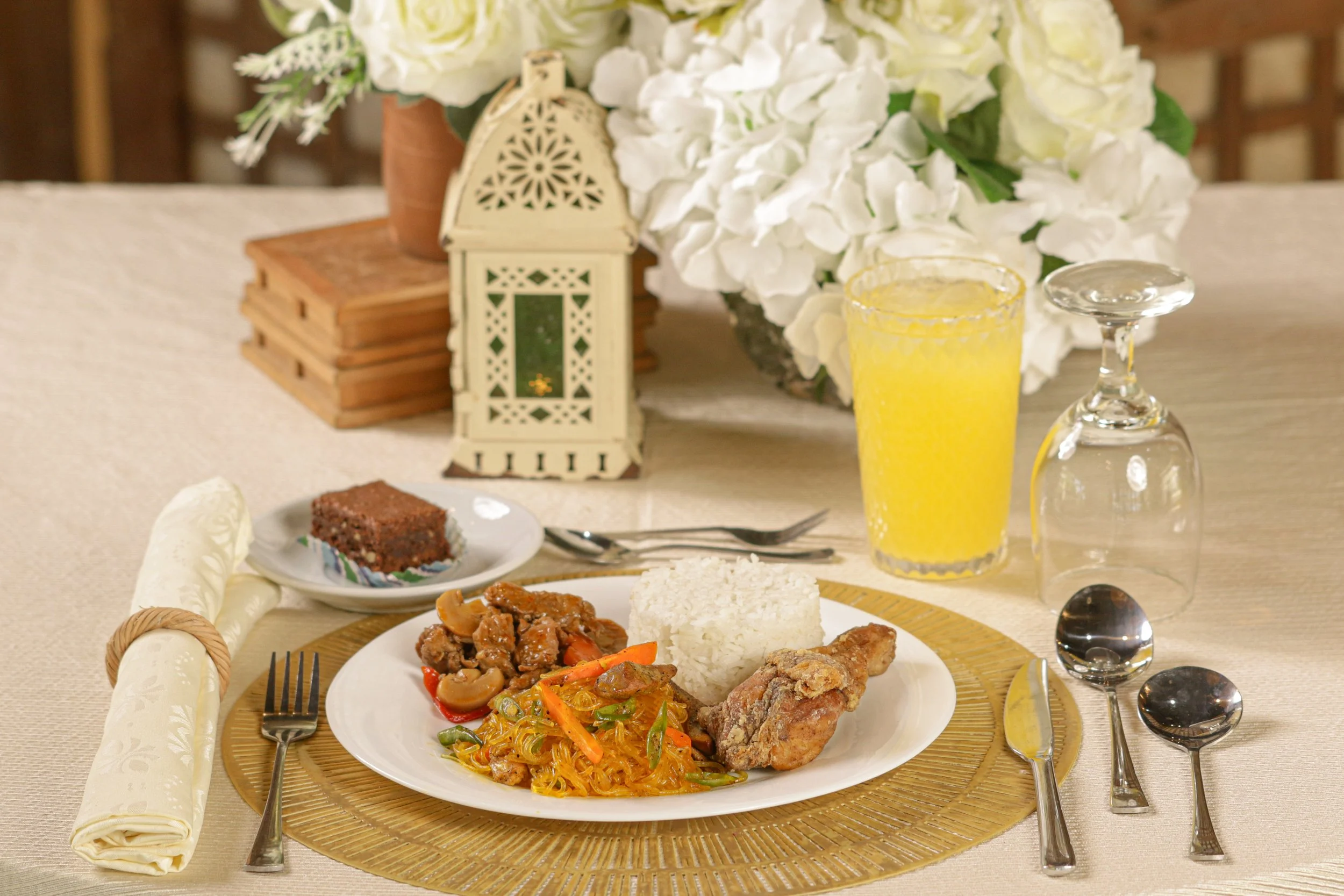 A dinner table with plated food including rice, stir-fried vegetables, beef, and fried chicken. There are two forks, two teaspoons, a knife, and a napkin on a beige tablecloth. A glass of yellow drink and an upside-down wine glass are on the table. In the background, there is a floral centerpiece with white flowers, a decorative lantern, and a stack of wooden boxes.