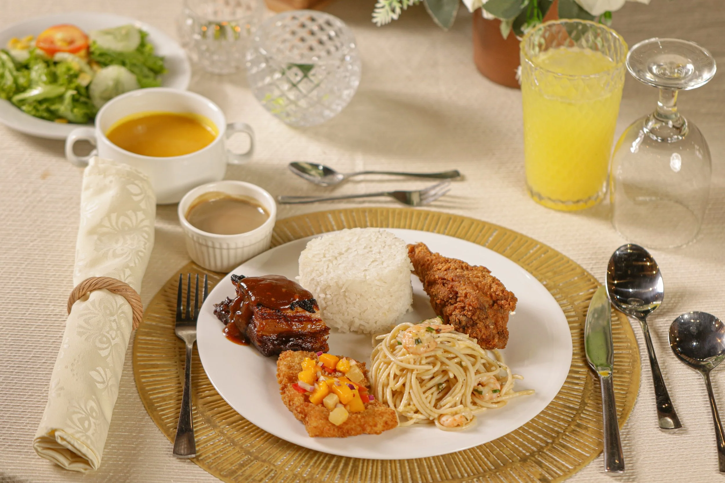 A plated meal of rice, fried chicken, shrimp pasta, ribs with sauce, and fried fish with fruit salsa on a decorative placemat. In the background, there is a salad, soup, and drinks including a yellow beverage and an empty glass. Silverware and napkin are arranged on a beige tablecloth.