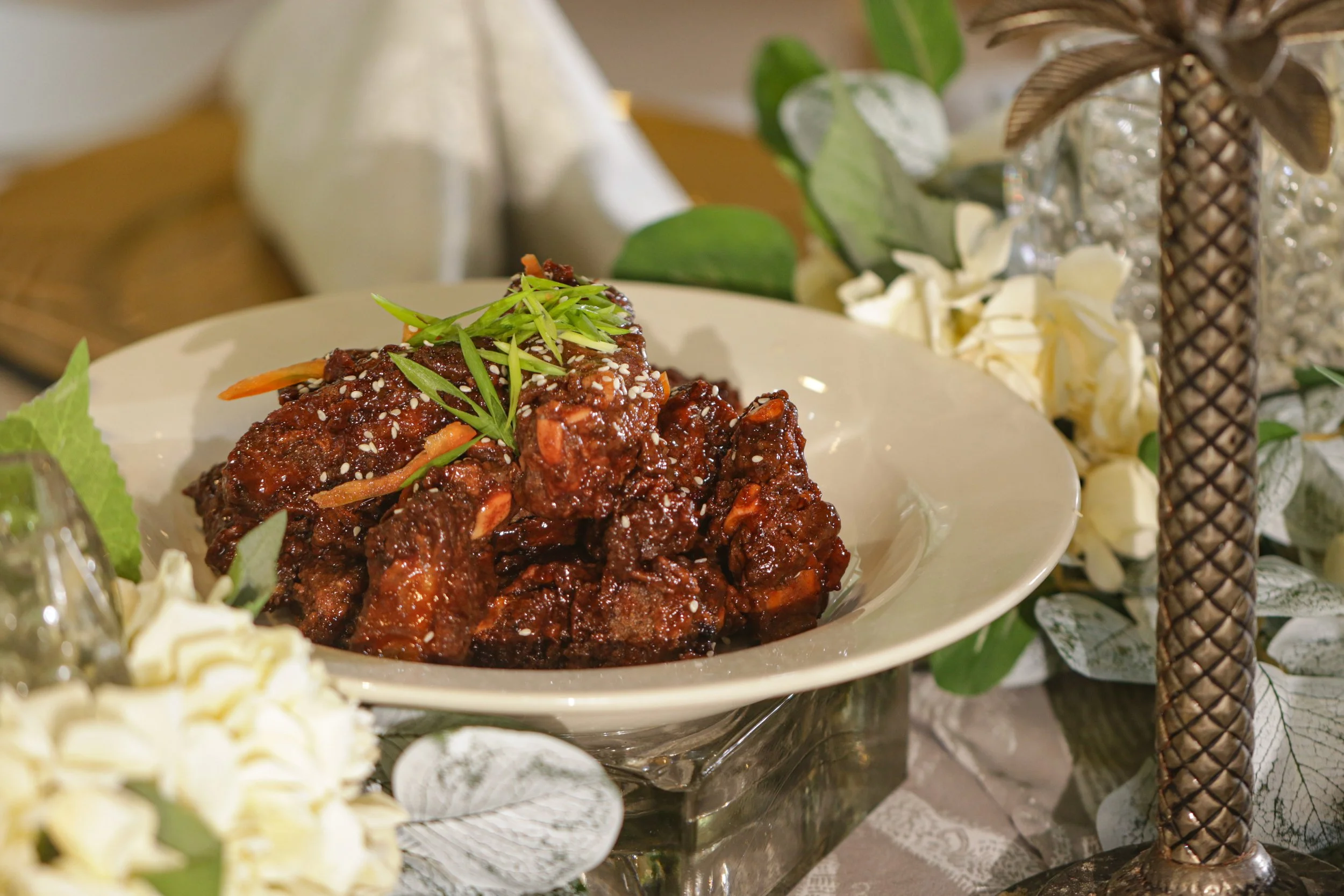 Sweet and sticky glazed beef or pork ribs garnished with green onions, served on a white plate with decorative flowers and greenery in the background.