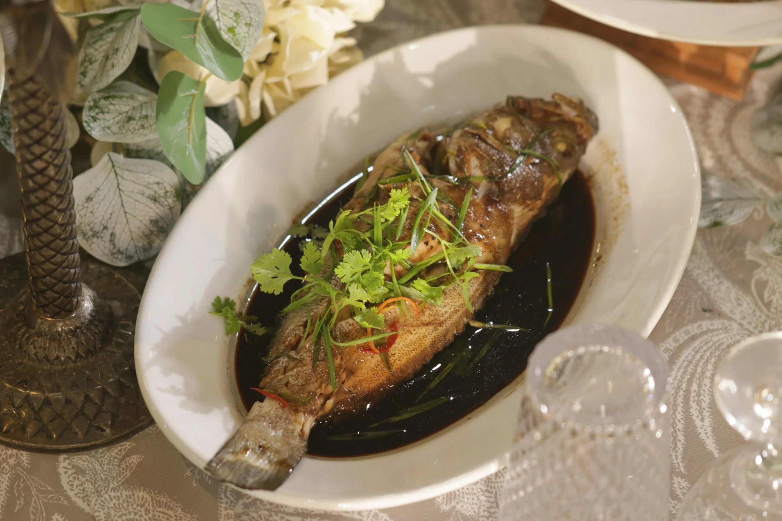 Whole fish cooked in dark soy sauce with cilantro and sliced chili on top, served in a white oval dish, with decorative foliage and crystal glassware on a patterned tablecloth.