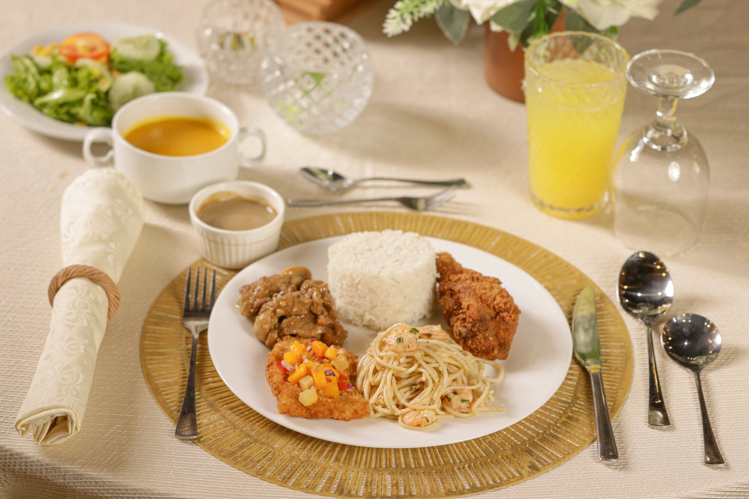 A fancy dinner table with a plate of rice, fried chicken, pasta, and meat, surrounded by salad, soup, drinks, and utensils.