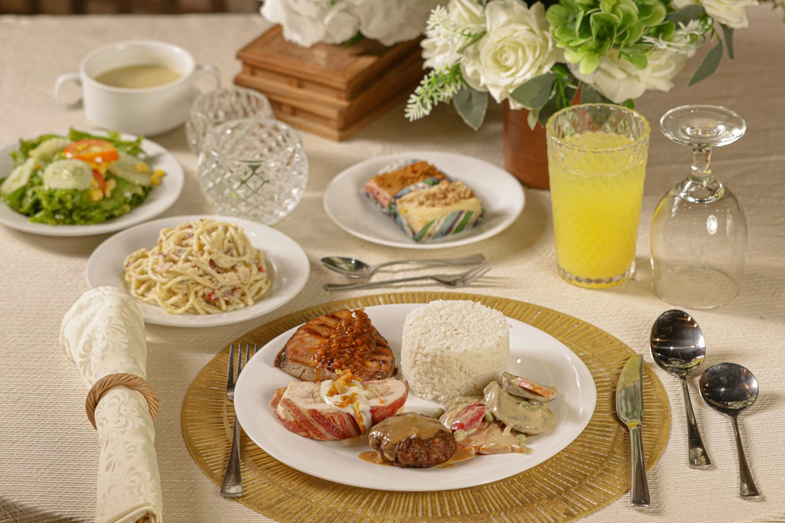 A formal dinner table set with a main plate of rice and assorted meats, side dishes including salad, pasta, and desserts, and drinks like yellow juice, with flowers and glassware in the background.