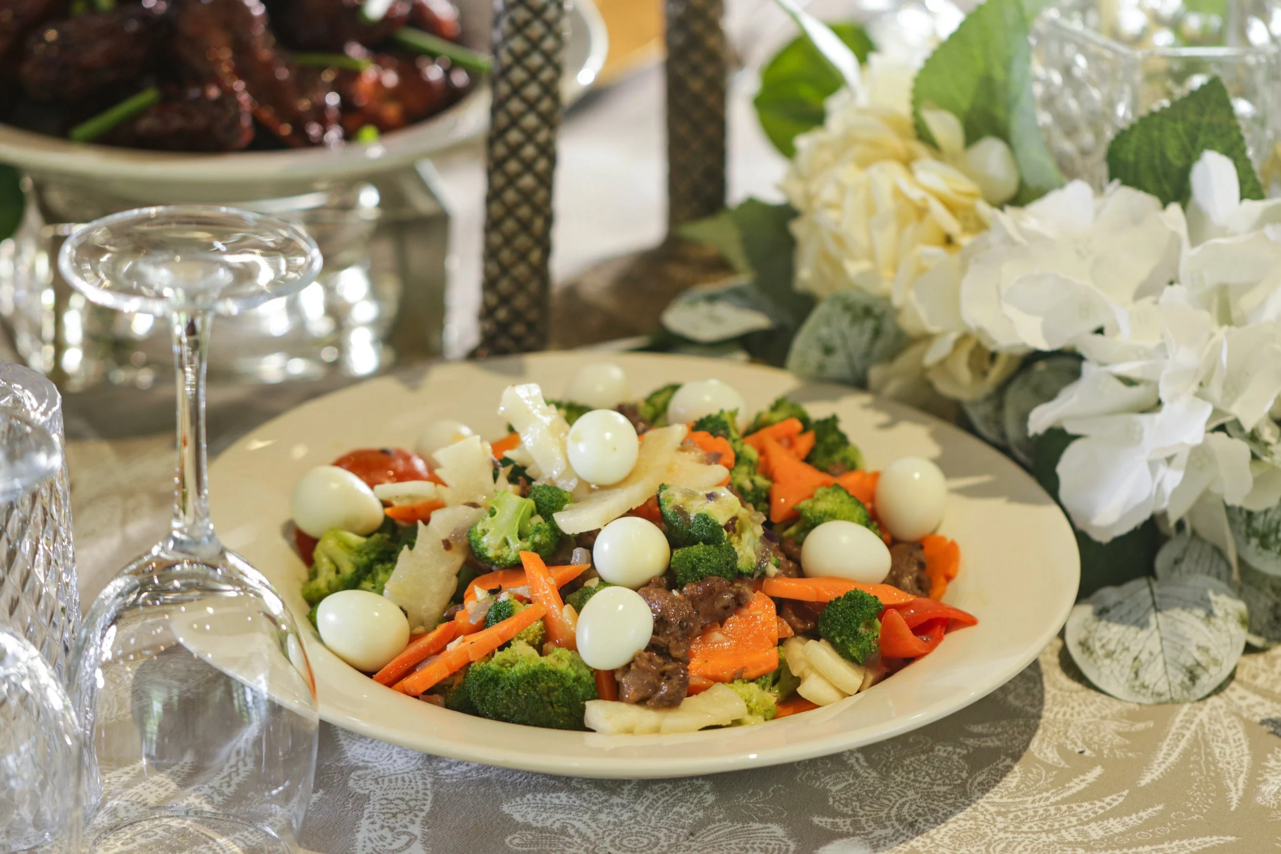 Colorful vegetable salad with quail eggs served on a white plate, part of an elegant table setting with floral decorations and glassware.