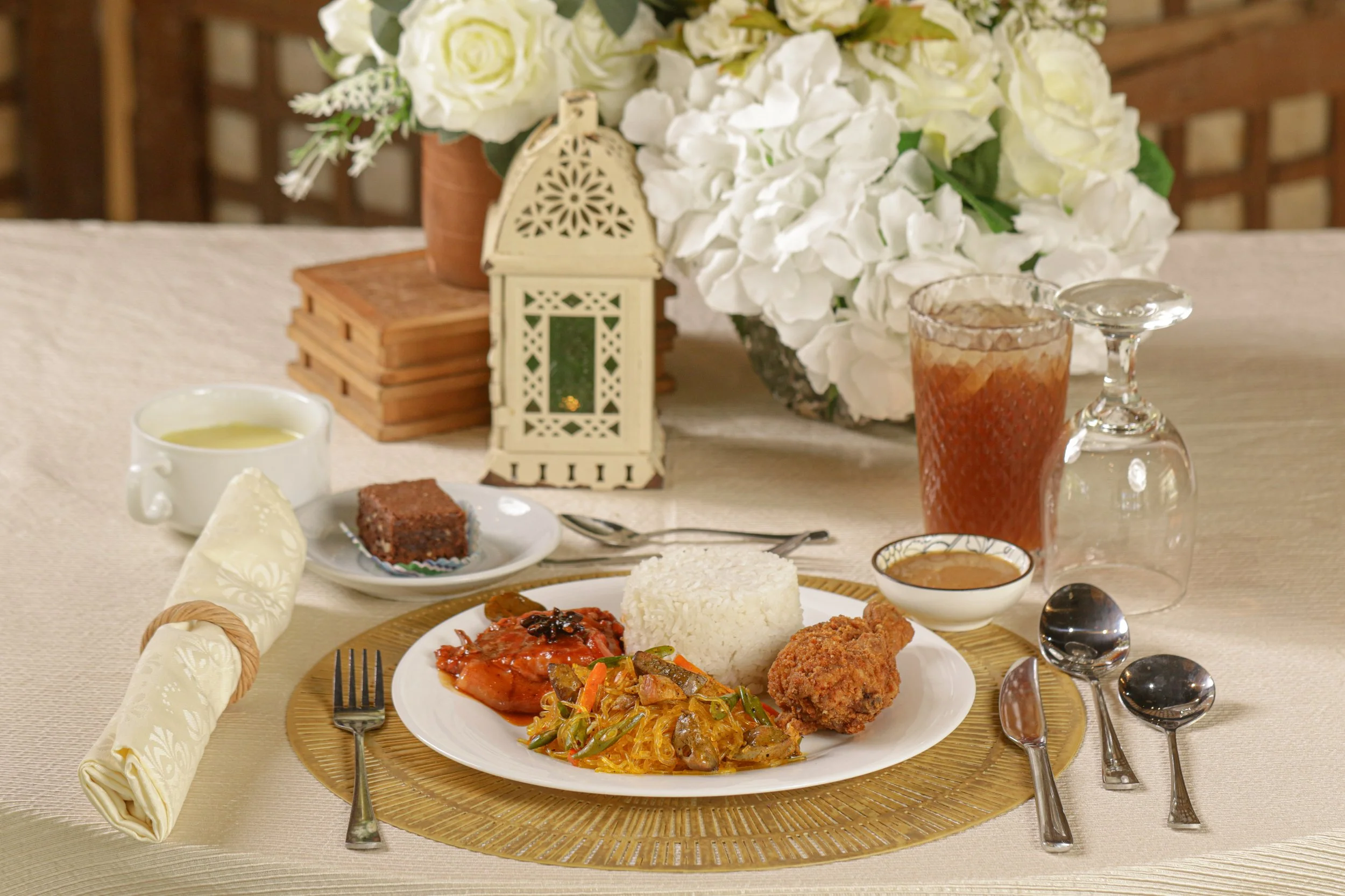 A formal table setting featuring a plate of rice, chicken, and vegetable dishes, along with side dishes and drinks. Decorated with a floral centerpiece and a lantern, on a cream tablecloth with utensils and napkin.