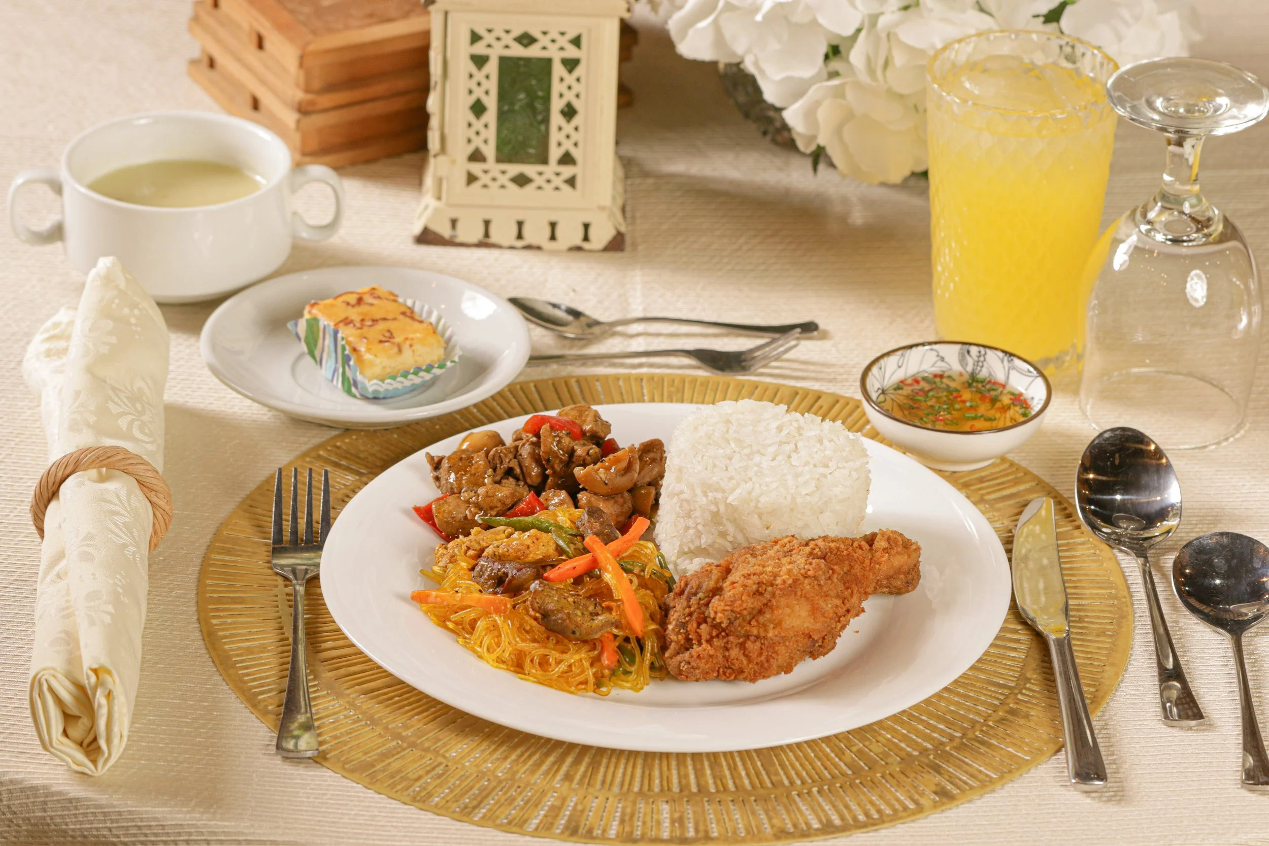 A plated meal with rice, fried chicken, noodles with vegetables, and a beef dish, along with a bowl of soup, a small pastry, and drinks including orange juice and a glass of water, on a table set with silverware and a golden placemat.