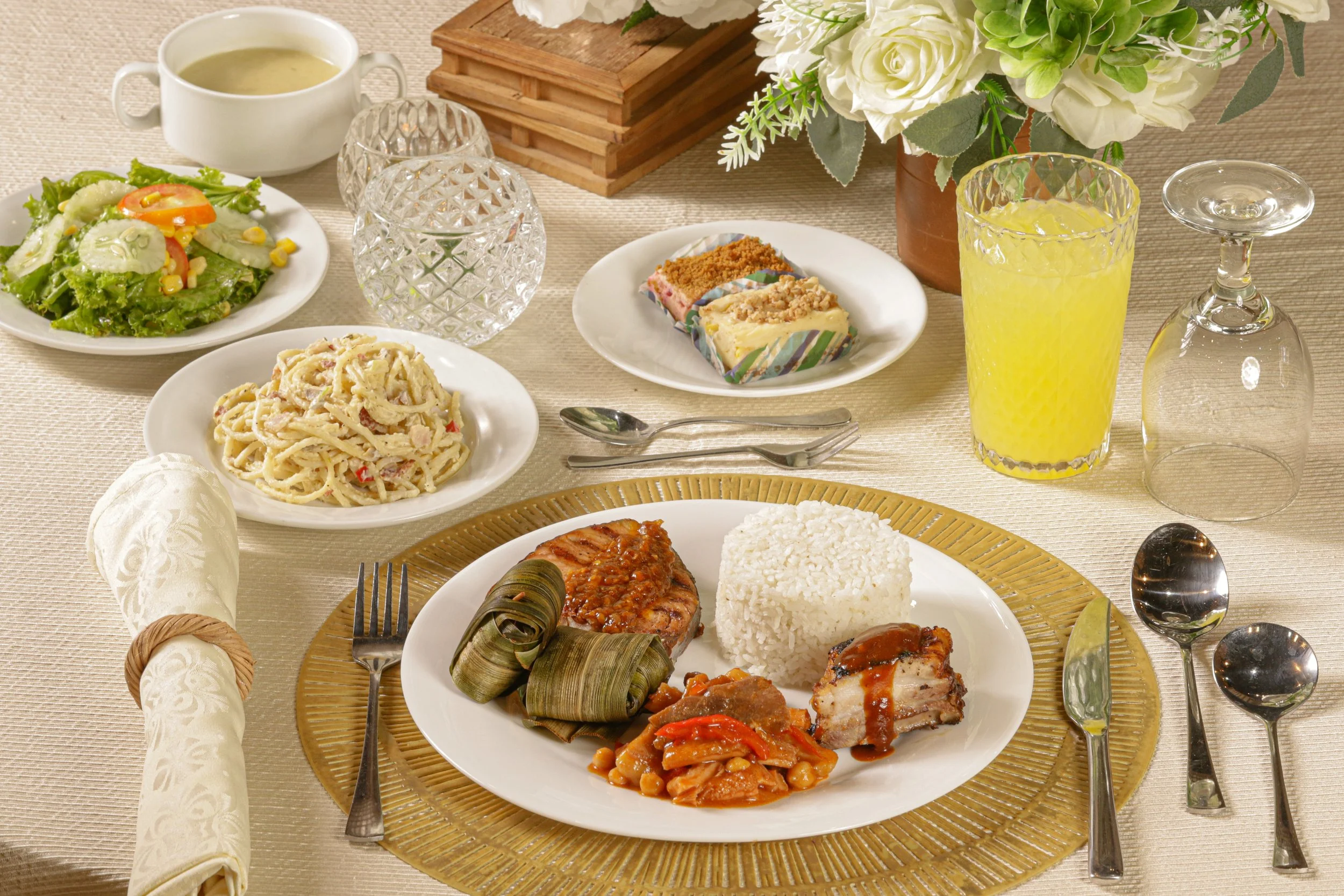 A formal dining table with a variety of dishes including a salad, pasta, and a main course with rice, chicken, and wrapped leaves; drinks including orange juice and water; and decorative flowers.