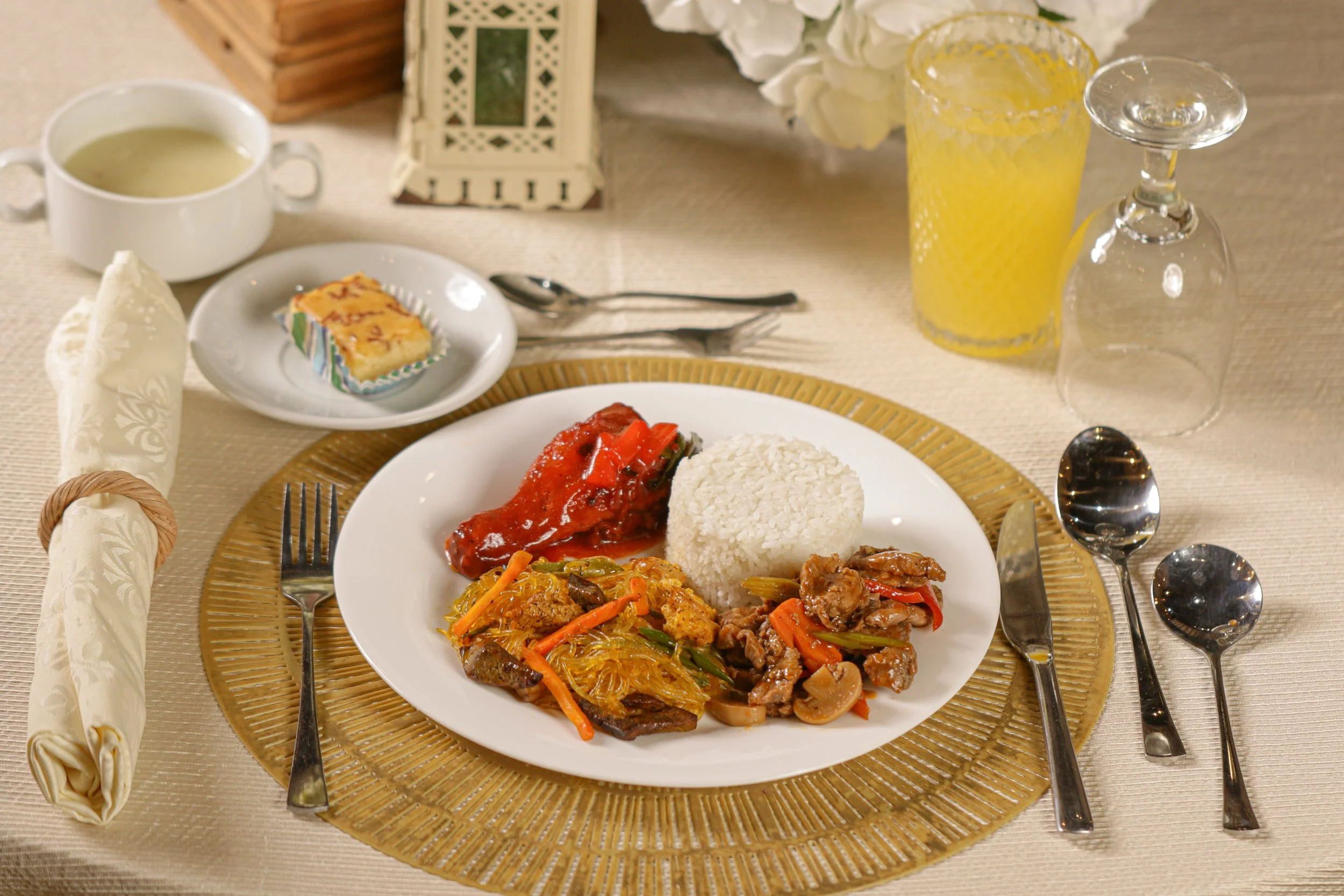 A dinner plate with rice, meat, and vegetables, on a gold-rimmed charger. Surrounding the plate are utensils, a glass of orange juice, a wine glass, a bowl of soup, a slice of cake, a rolled napkin, and a fork. In the background, there are flowers, a lantern, and additional tableware.
