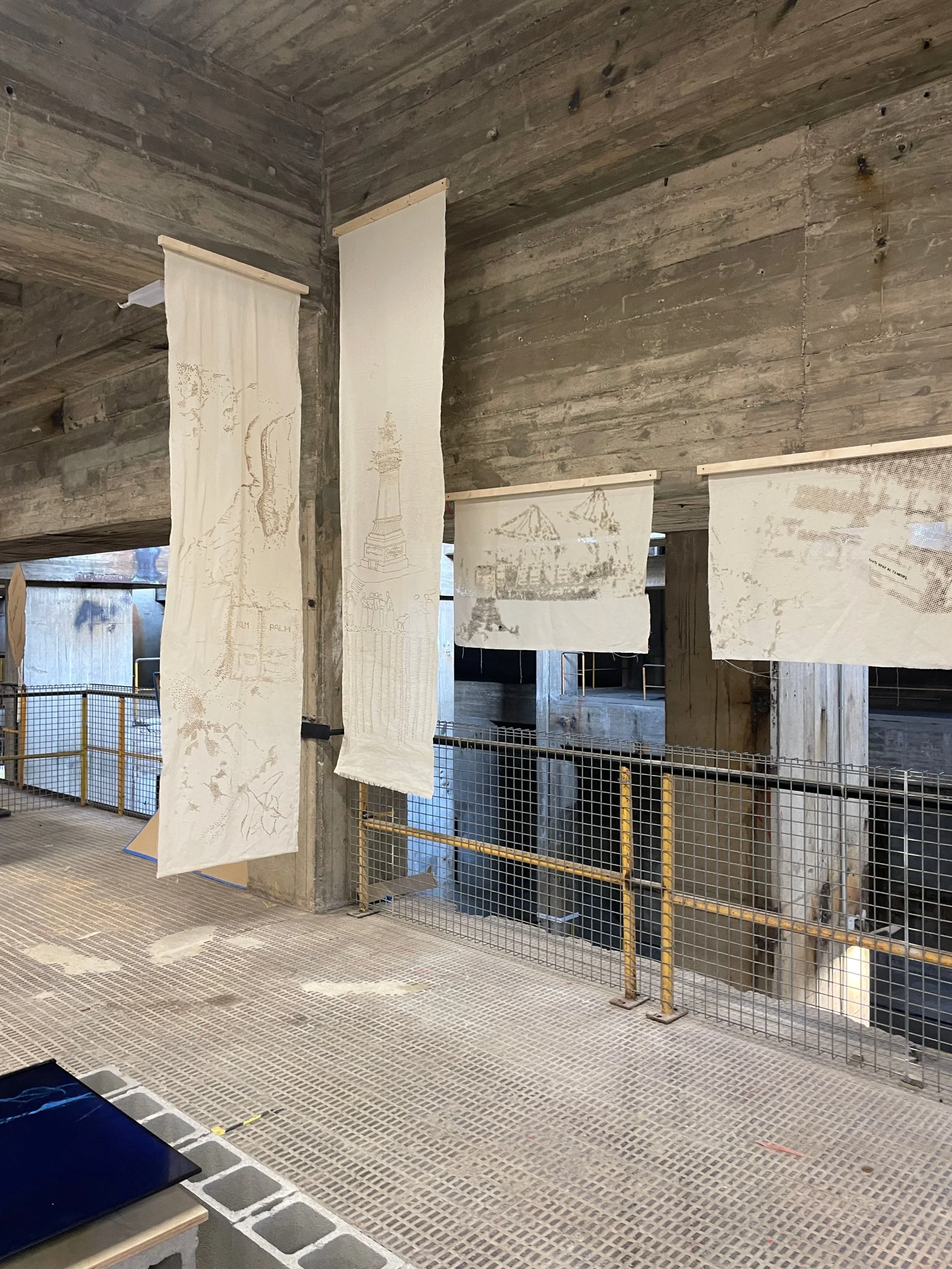 Tall calico banners from the Revised Cartographies workshop, one showing a burnt drawing of a monument, hanging in a concrete hall at the UIA World Congress of Architects 2026, Barcelona.