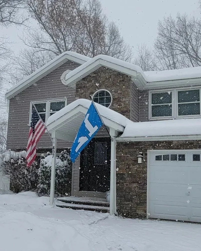 A two-story house with a brick facade and vinyl siding, covered in snow, with American and Tesla flags displayed near the front door.