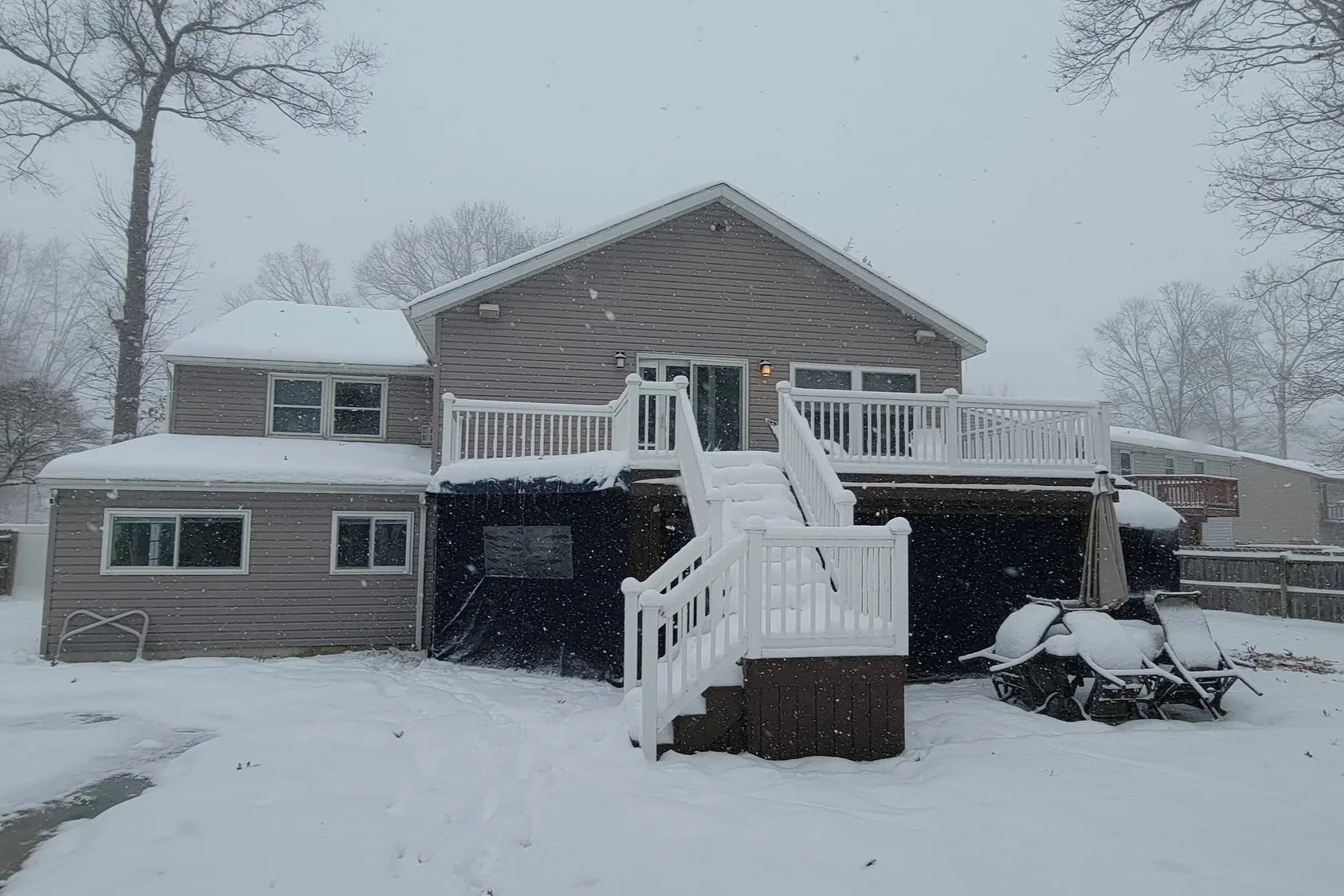 Back of a house during snowfall with steps leading to a deck, covered patio furniture, and snow-covered yard and trees.