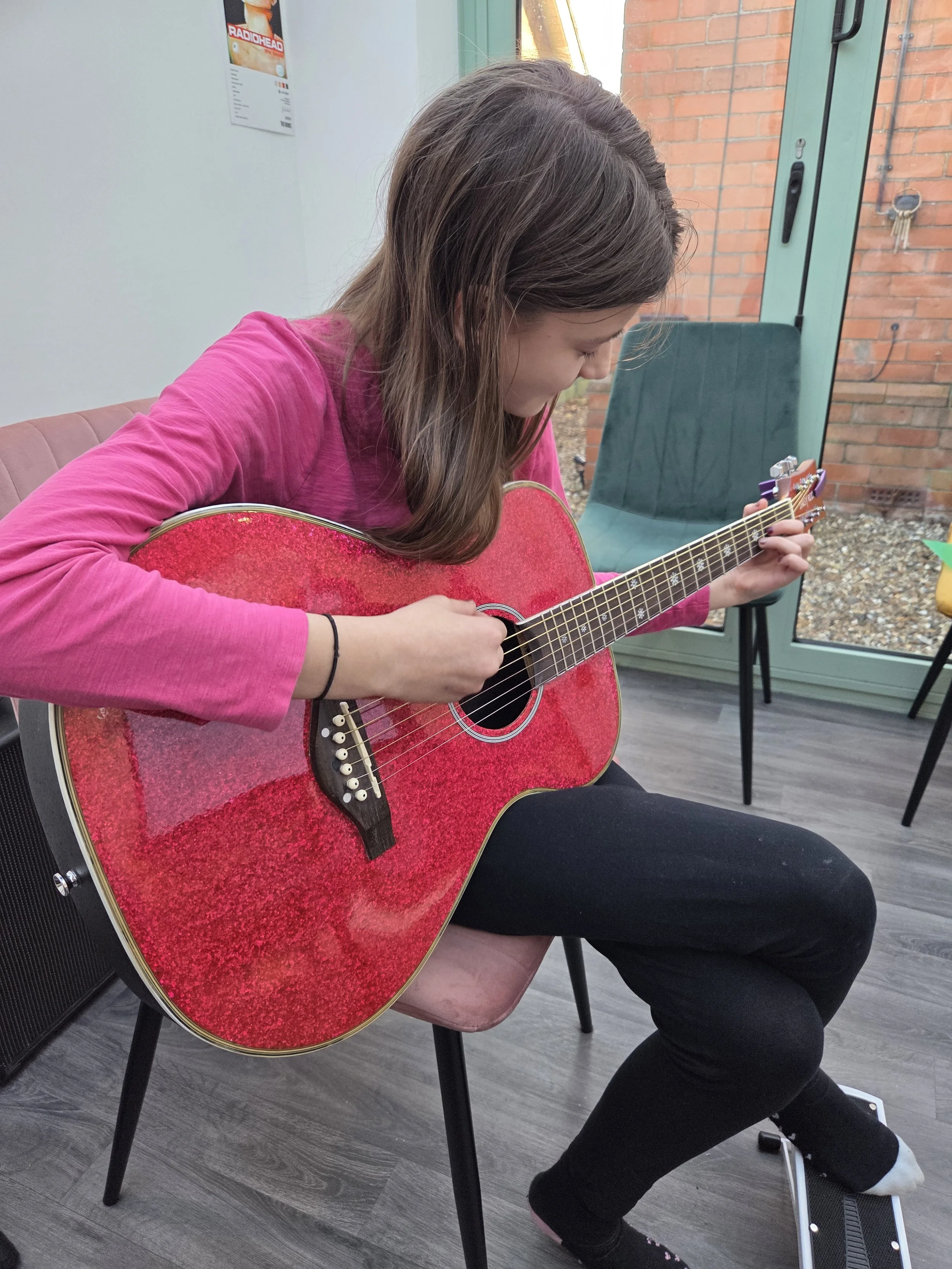A young woman with long brown hair wearing a pink long-sleeve top playing a red acoustic guitar while sitting on a pink chair indoors near a glass door.