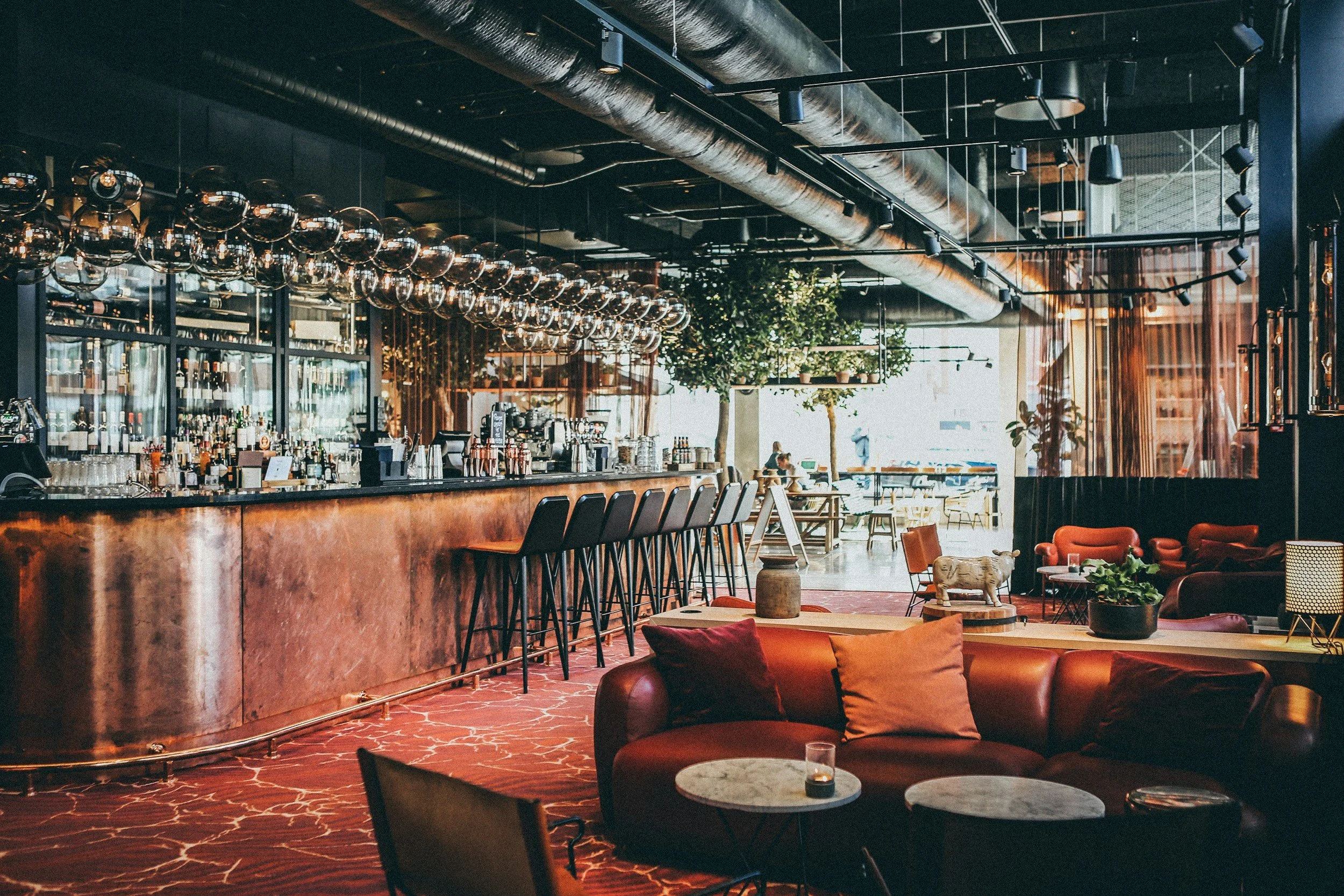 Interior of a modern bar and lounge area with a curved copper bar counter, high black chairs, and warm lighting, featuring a red patterned carpet, plush seating, potted plants, and large windows letting in natural light.