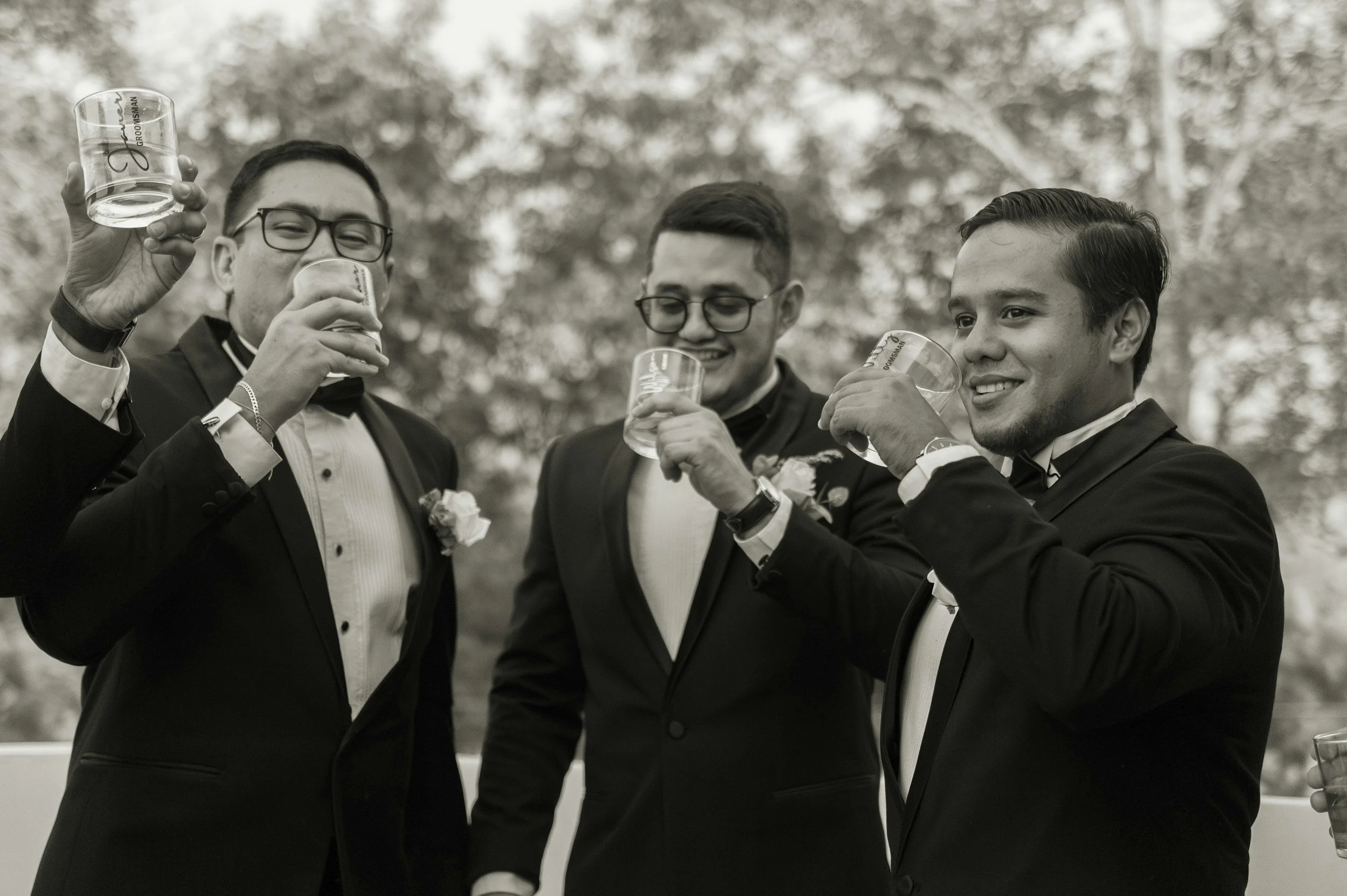 Three men in tuxedos celebrating with drinks outdoors, smiling and raising glasses