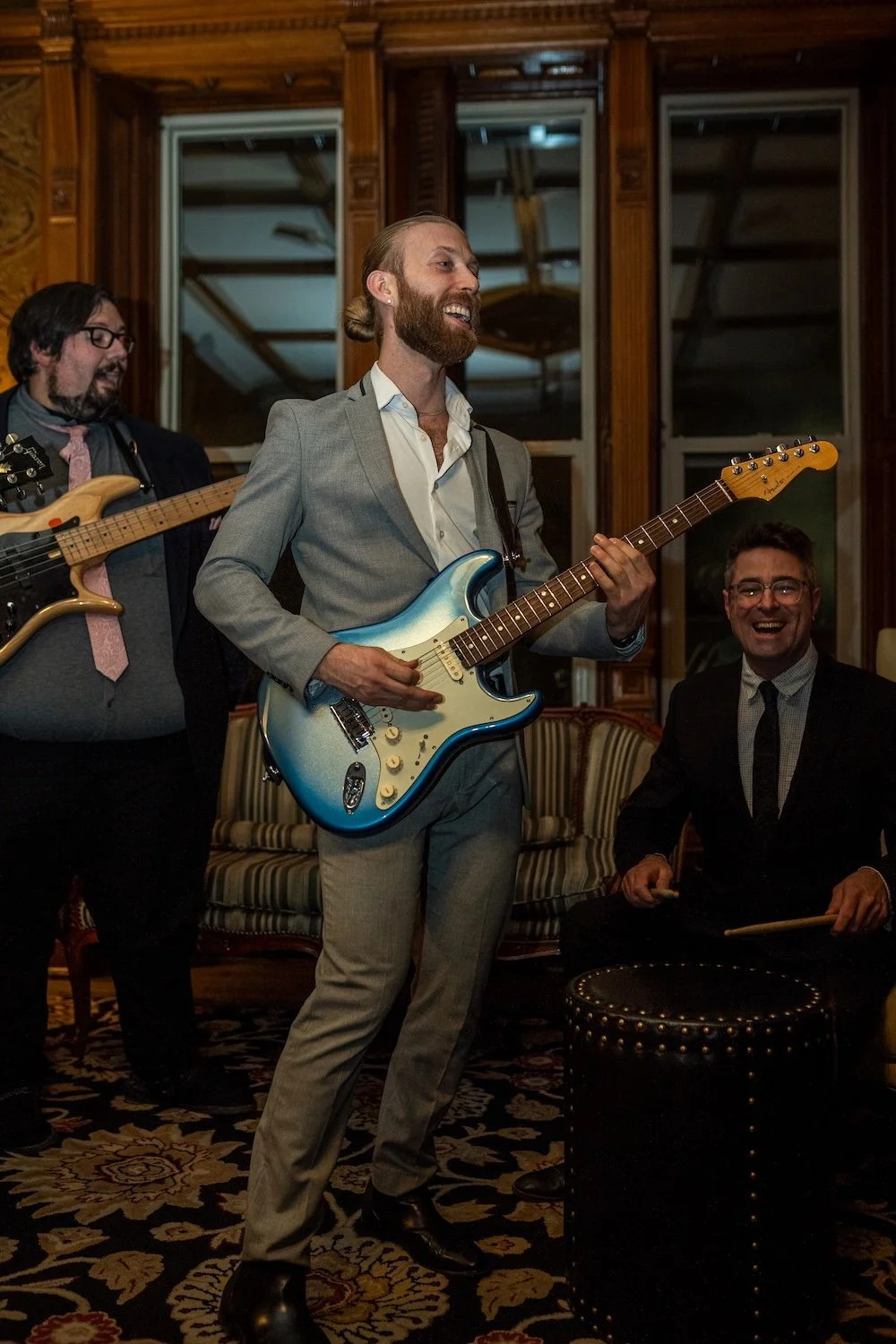 Three men playing musical instruments at a social gathering in a warmly lit room with wooden walls and large windows.