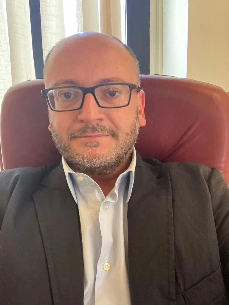 Uomo con occhiali, camicia bianca e giacca scura, seduto su una sedia di pelle rossa, sullo sfondo tende chiare vicino a una finestra