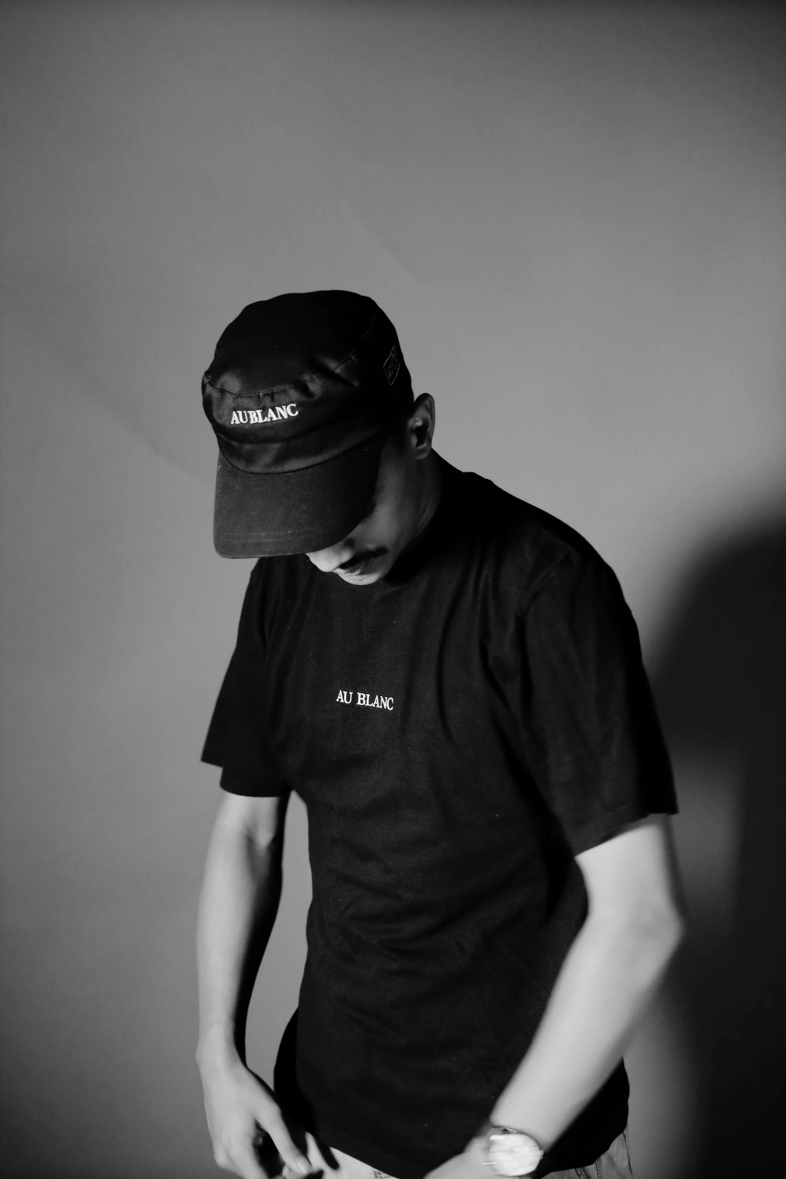 A young man dressed in black, wearing a cap and a t-shirt with the words 'AU BLANC,' looking downward against a plain background.