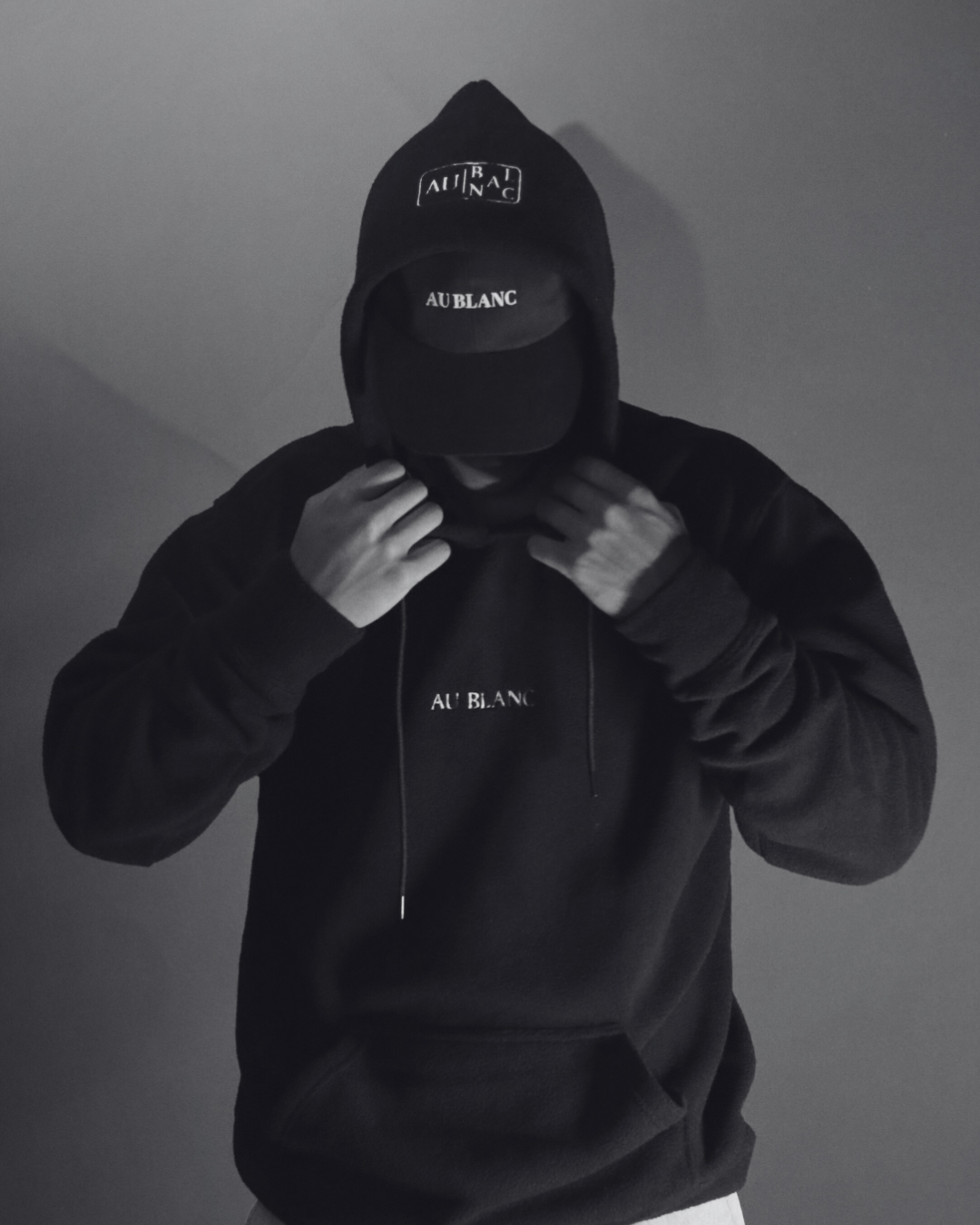 A person dressed in black, wearing a hoodie, cap, and a mask, with the words "AUB BLANC" written on all items, standing against a gray wall.