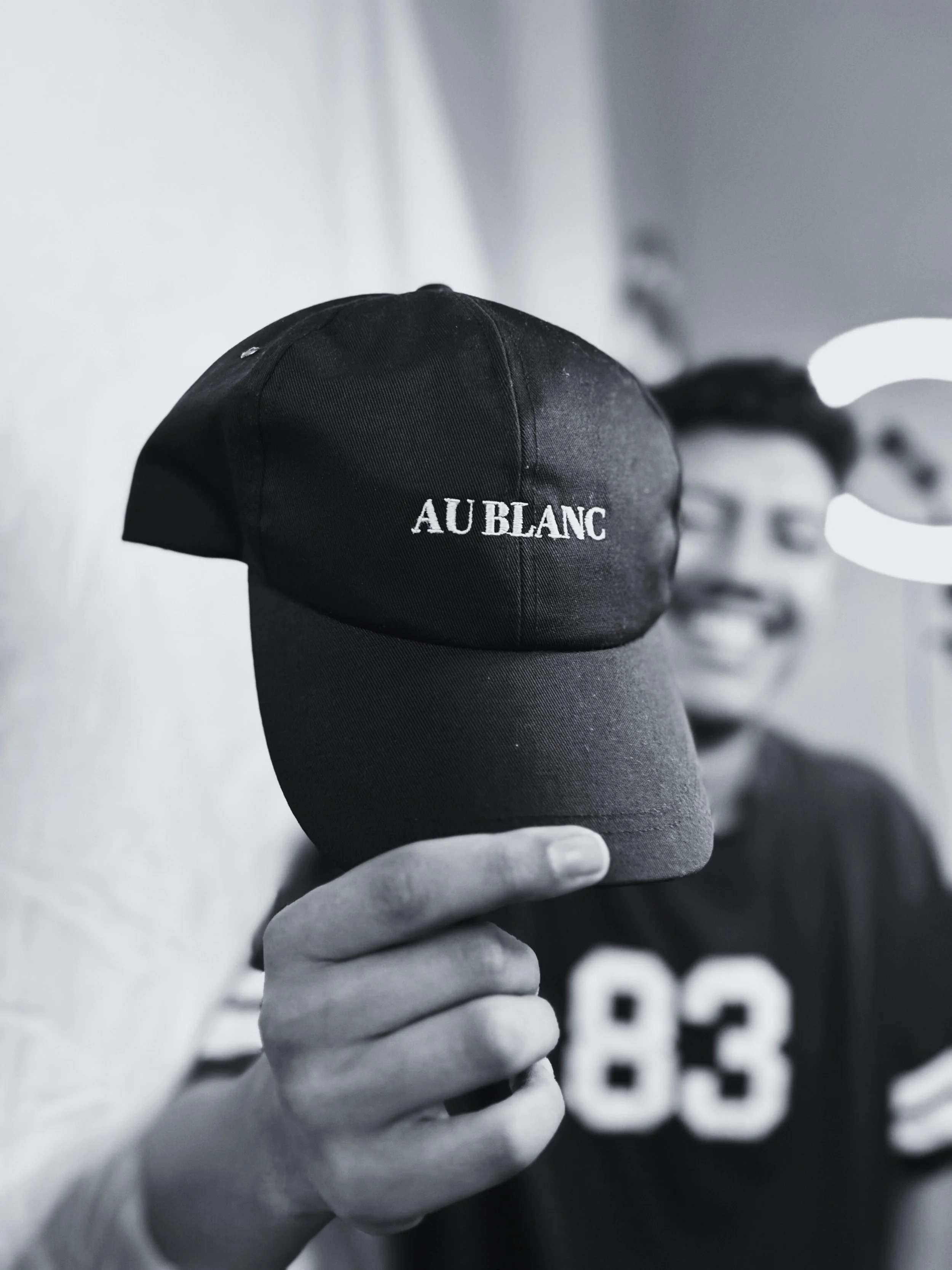 Person holding a black baseball cap with the words 'AU BLANC' embroidered on it, in front of a blurred smiling face in the background.