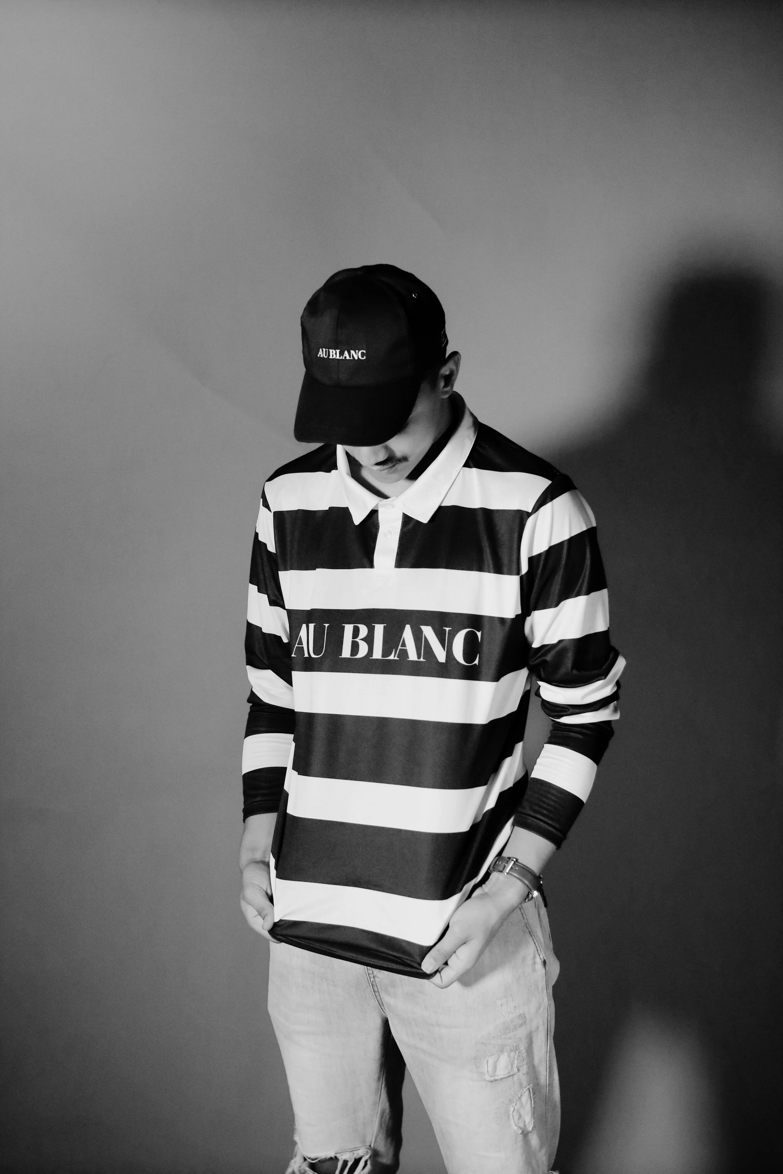Black and white photo of a person wearing a striped long-sleeve shirt with 'AU BLANC' written on it, a black cap with the same text, and light-colored ripped jeans, standing against a plain wall.