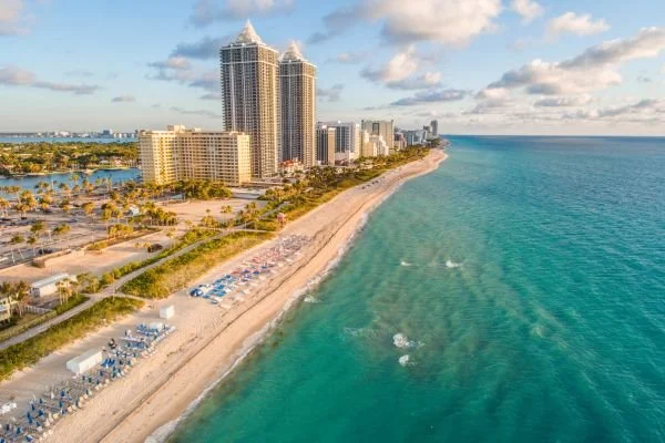 Aerial view of a beach with skyscrapers along the shoreline, clear blue ocean, and a partly cloudy sky.