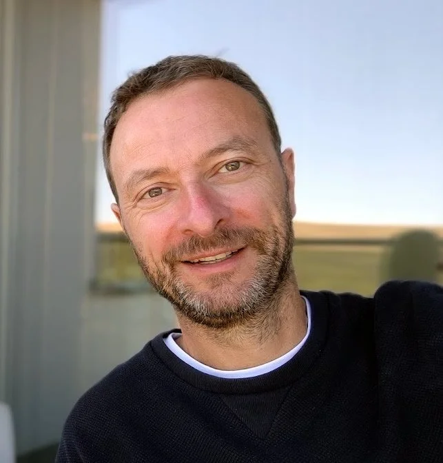 A smiling man with light brown hair and a beard taking a selfie outdoors, with a clear sky and field in the background.