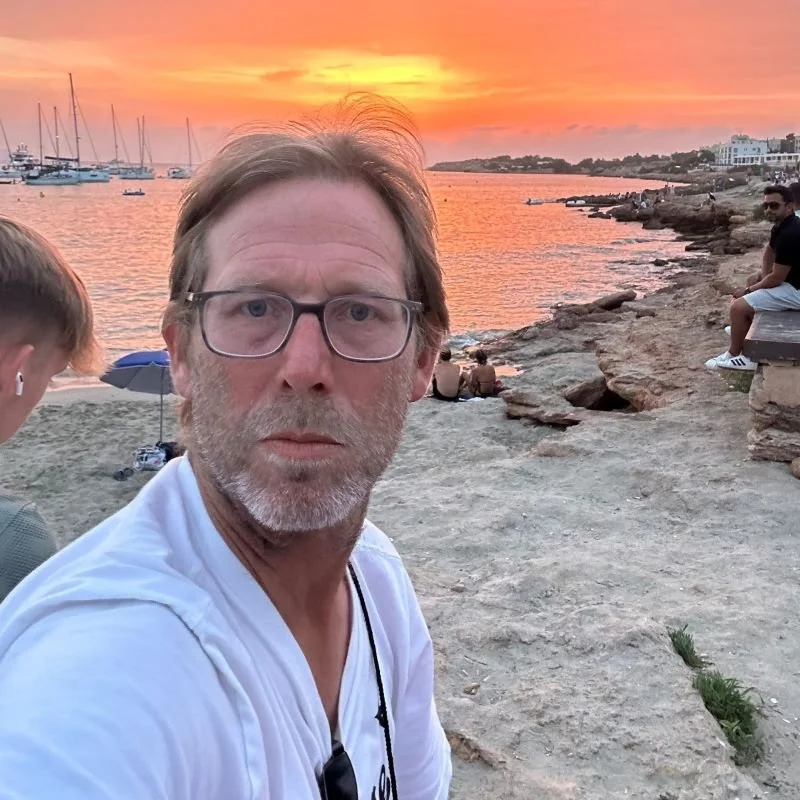 A man with glasses and a beard taking a selfie near a body of water during sunset, with boats anchored in the water and a rocky shoreline with other people in the background.
