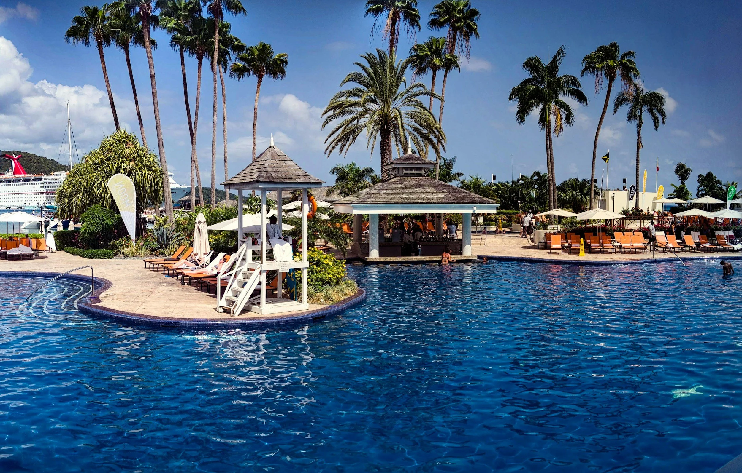 Poolside scene at a tropical resort with lounge chairs, umbrellas, palm trees, and a large cruise ship in the background.