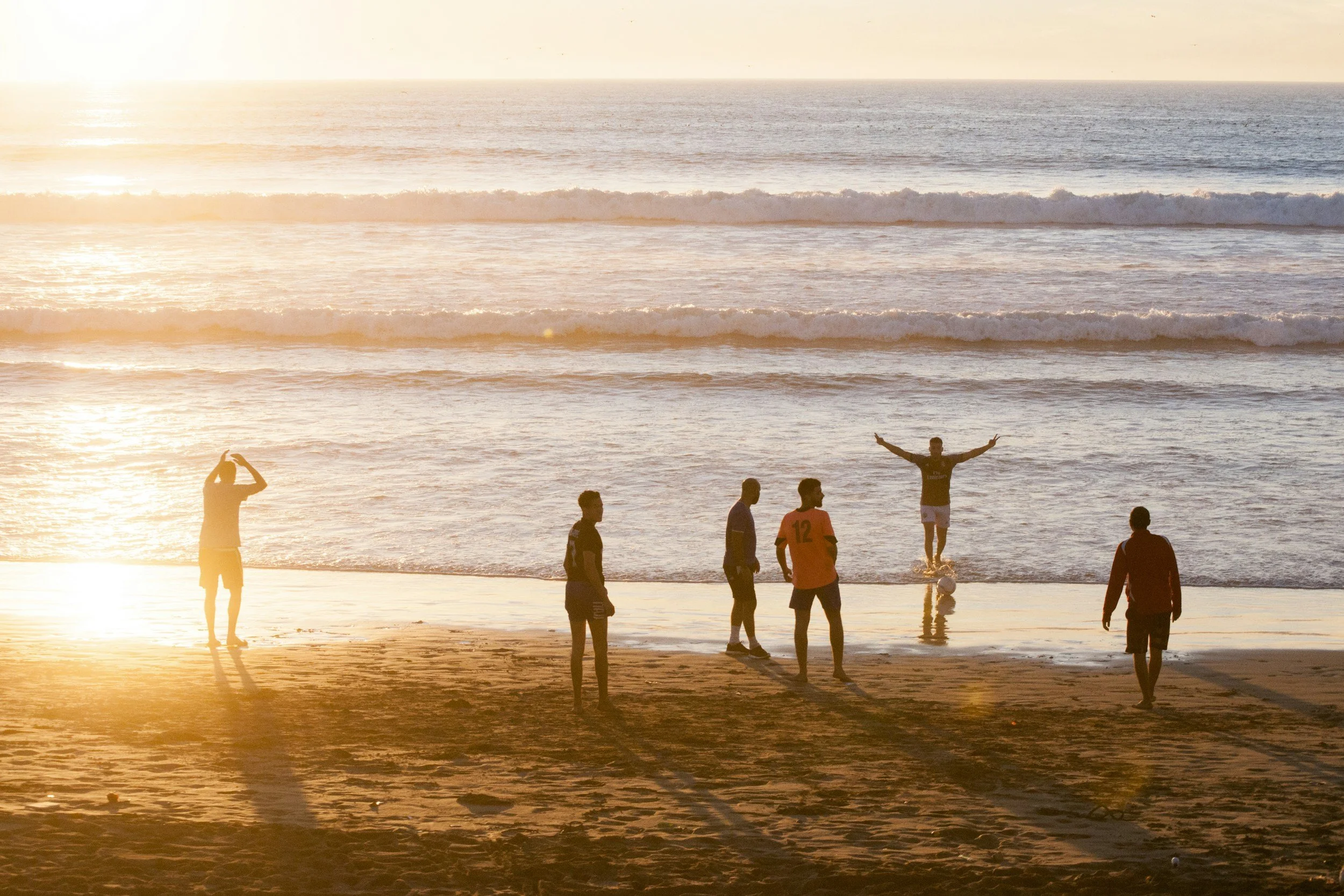 A group of six people on a beach playing soccer during sunset; one person is standing in shallow water with arms outstretched, while others are on the sand, and one person is taking a photo.
