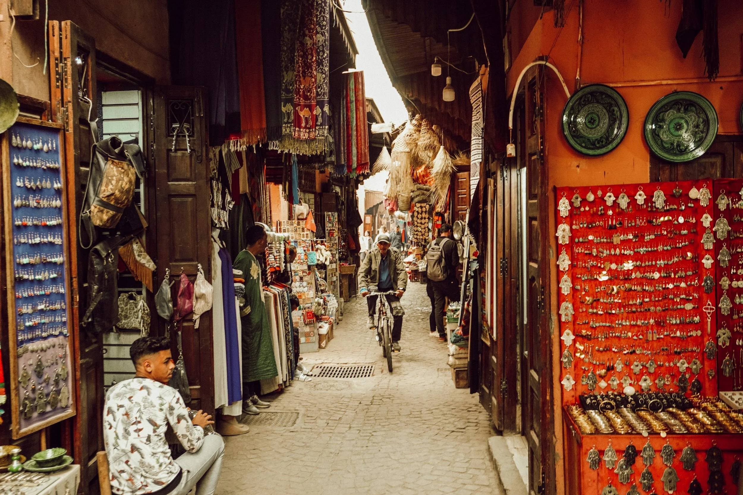 A busy marketplace with vendors selling jewelry, textiles, and souvenirs. People shopping and walking through a narrow alley with colorful displays and hanging fabrics.