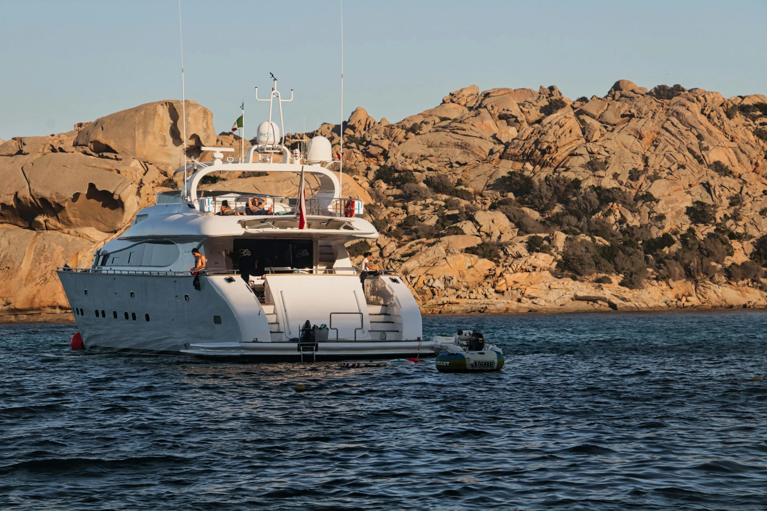 A white luxury yacht anchored near rocky coastline with passengers relaxing on deck and a small boat attached nearby.