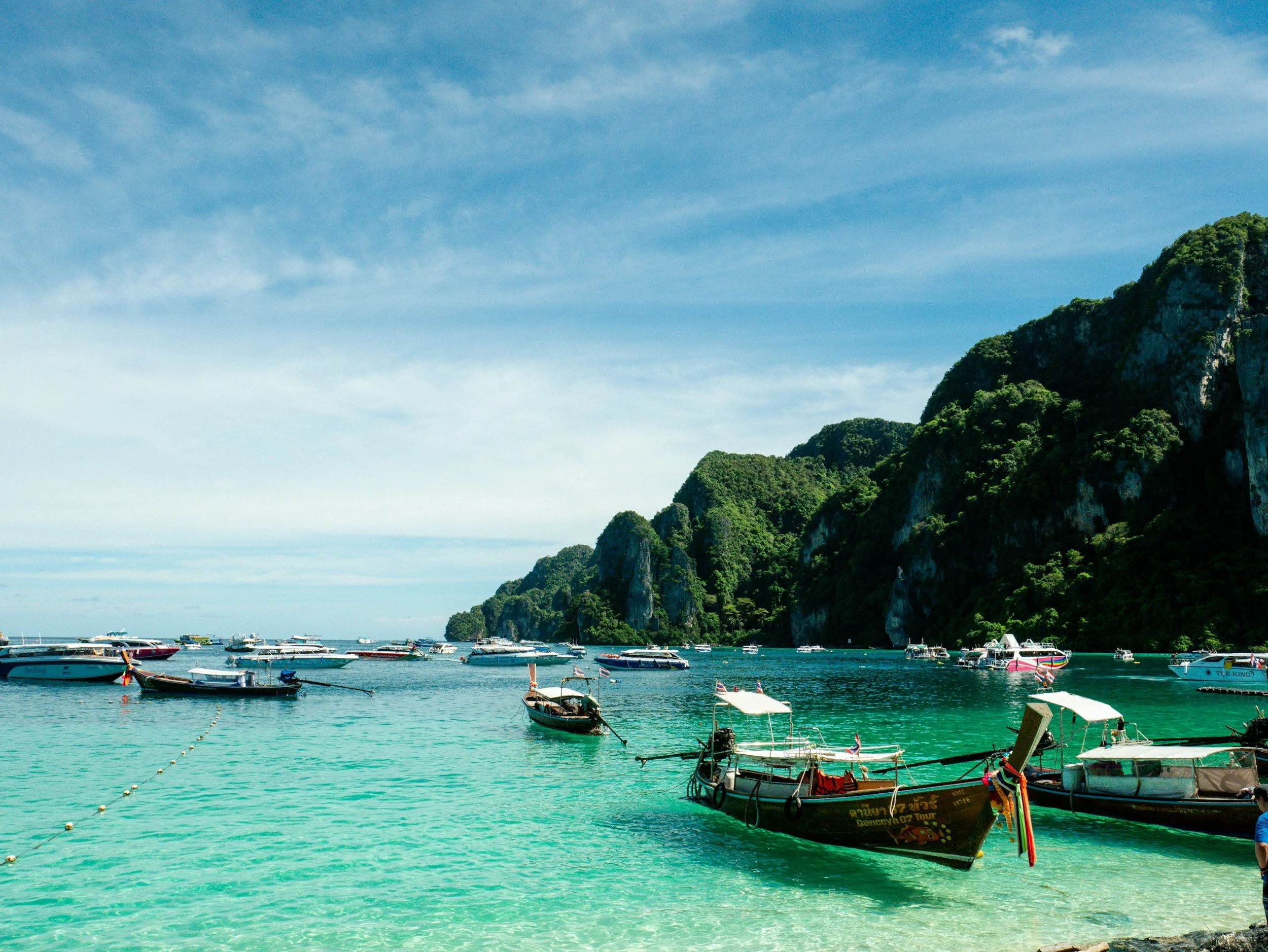 A tropical beach with clear turquoise water and multiple boats anchored near the shore, against green hills and a blue sky with some clouds.