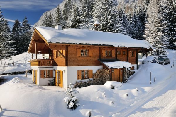 A wooden cabin with a snow-covered roof in a snowy forest landscape.