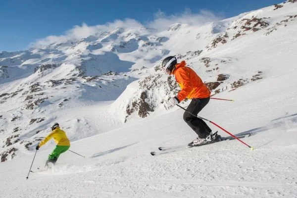 Two skiers in bright jackets skiing down a snowy mountain with a mountain range in the background.
