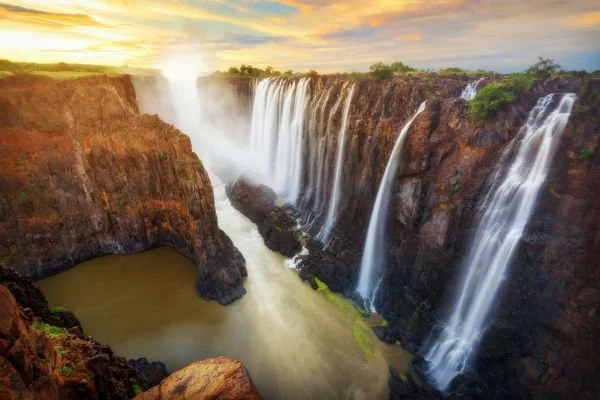A wide view of a large waterfall flowing into a river with cliffs on either side during sunset.