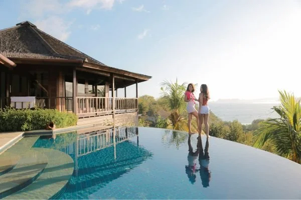 Two women in swimsuits talking by an infinity pool at sunset, with a tropical landscape and ocean view in the background.