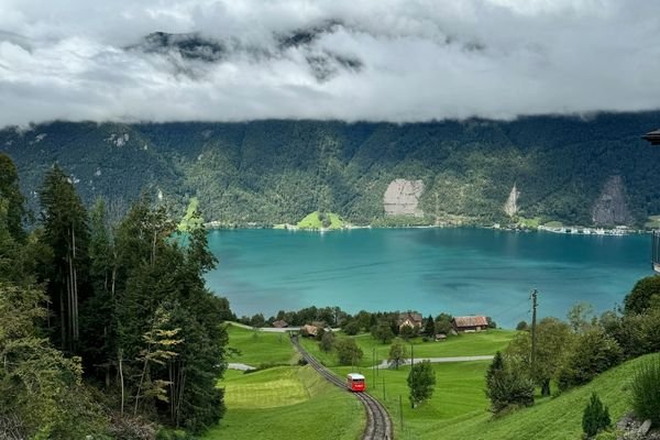 A scenic view of a lake surrounded by lush green hills and forested mountains under a cloudy sky, with a red train on a track through the valley.
