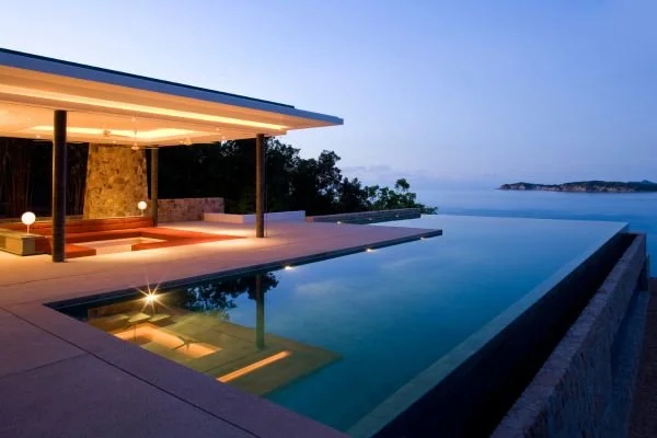 Modern house with a flat roof and an outdoor pool on a cliff overlooking the ocean at dusk.