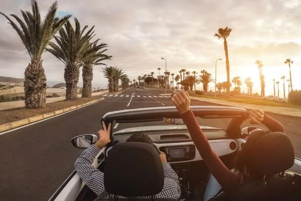 Two children in a convertible car driving along a road lined with palm trees at sunset.