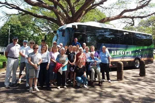 Group of senior adults standing and sitting in front of a large tour bus with a green and blue design that says 'Aurora Cest Traveler,' under a large tree on a sunny day.