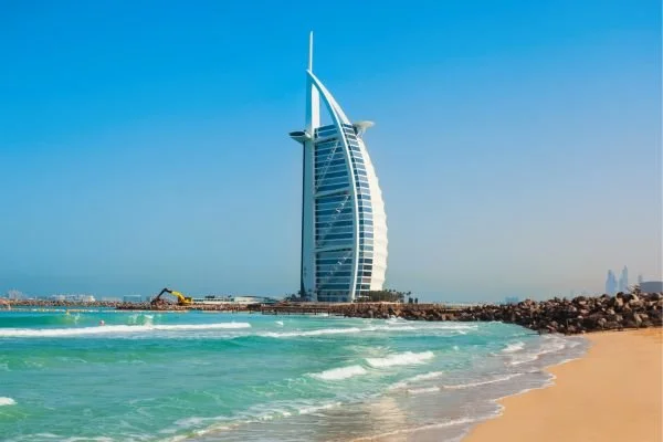 The Burj Al Arab hotel standing on the coastline with a sandy beach and waves in the foreground under a clear blue sky.