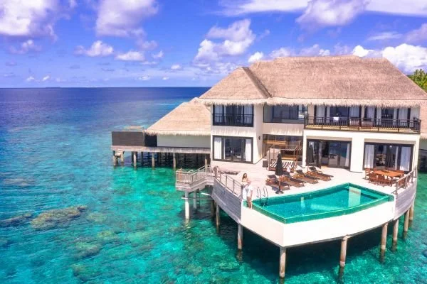 Overwater bungalow with a thatched roof, outdoor lounge chairs, and a private pool, situated above clear turquoise water.
