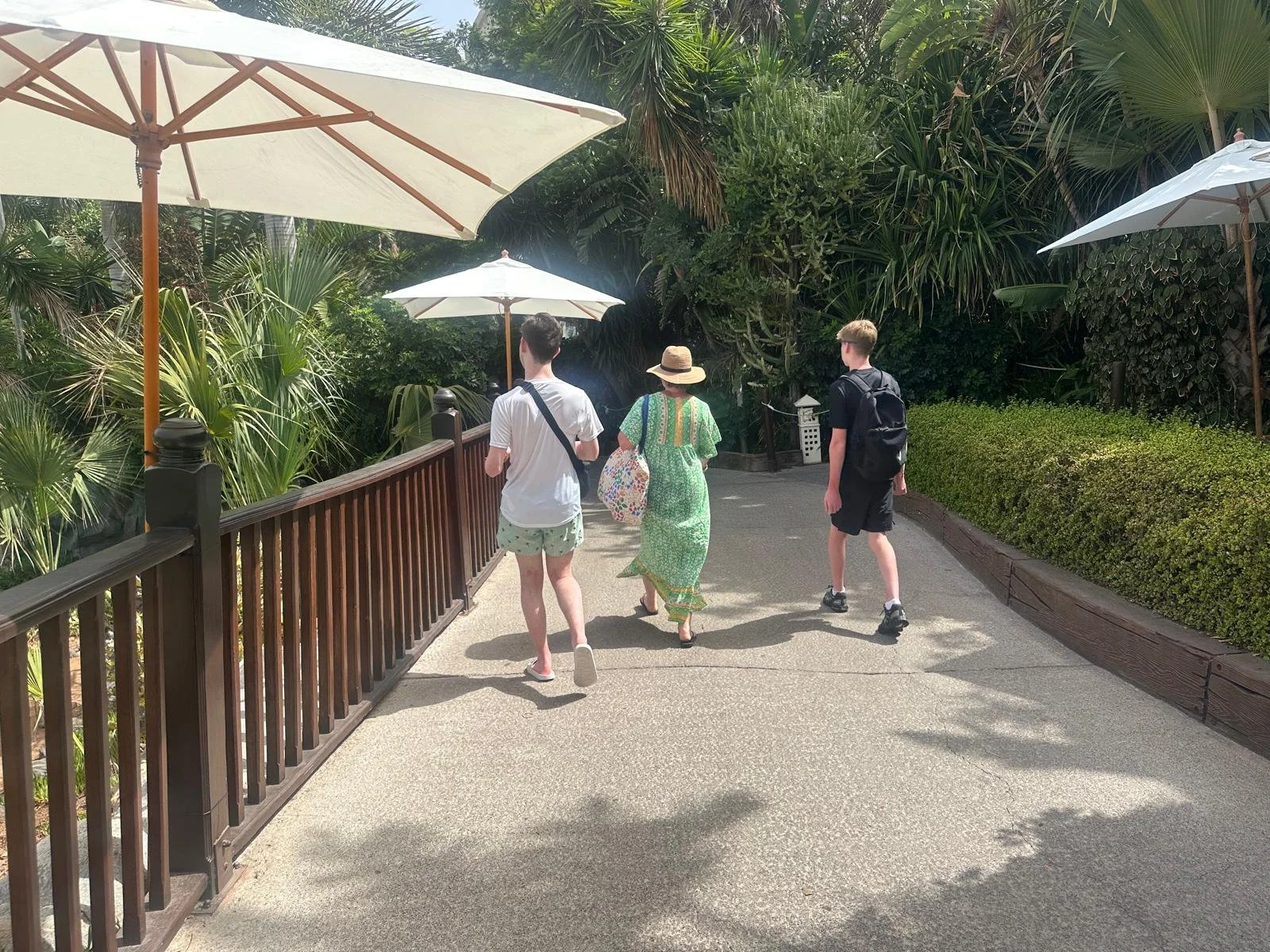 Three people walking on a garden pathway with lush greenery and white umbrellas overhead.
