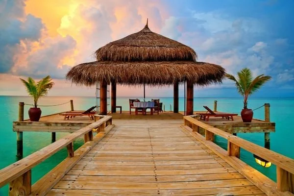 A wooden pier extending over turquoise water, leading to a seaside hut with a thatched roof, surrounded by two potted palm trees, at sunset.