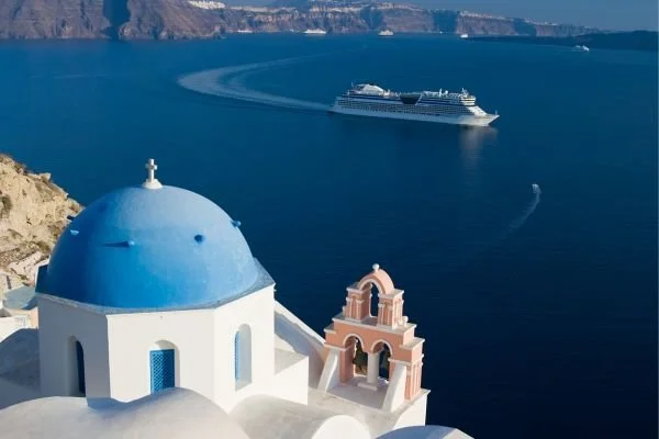 A white building with a blue dome and small bell tower overlooking the sea, with a large cruise ship sailing in the distance.