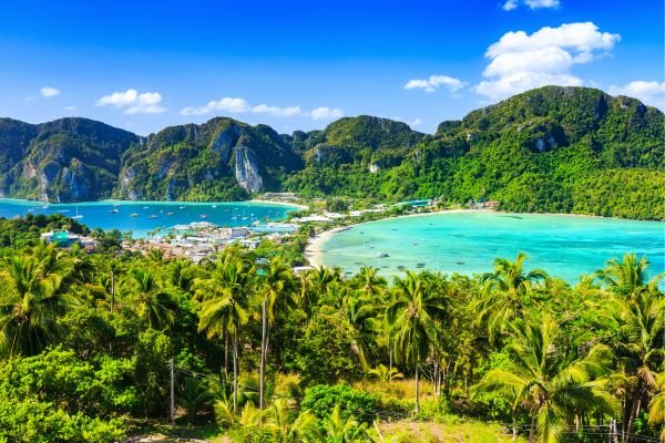 Tropical coastal landscape with lush green palm trees, clear blue waters, and mountainous islands in the background under a partly cloudy sky.