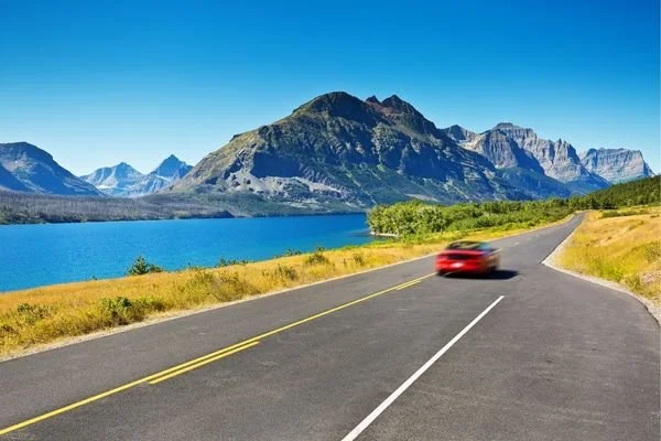 A red vehicle driving on a winding road next to a large blue lake with green shrubbery and mountains in the background under a clear blue sky.