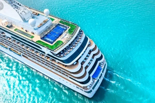 Aerial view of a cruise ship on the ocean, showing decks with a swimming pool, sports courts, and outdoor seating areas.