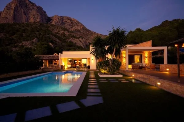 Modern house with a swimming pool, outdoor seating area, and landscaping, set against a backdrop of mountains at dusk.