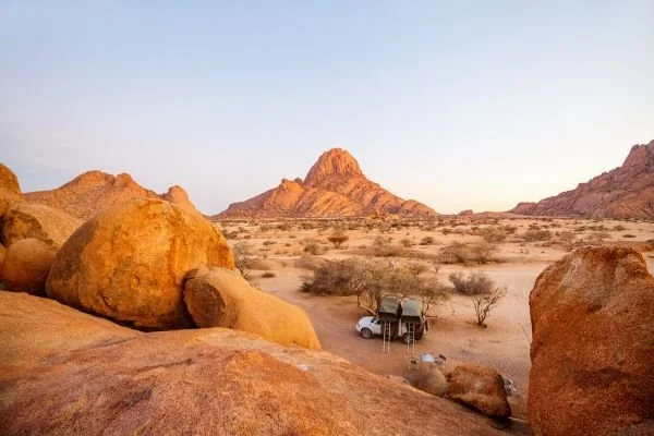 Desert landscape with large rocks, sparse vegetation, and a vehicle parked near the horizon under a clear sky.